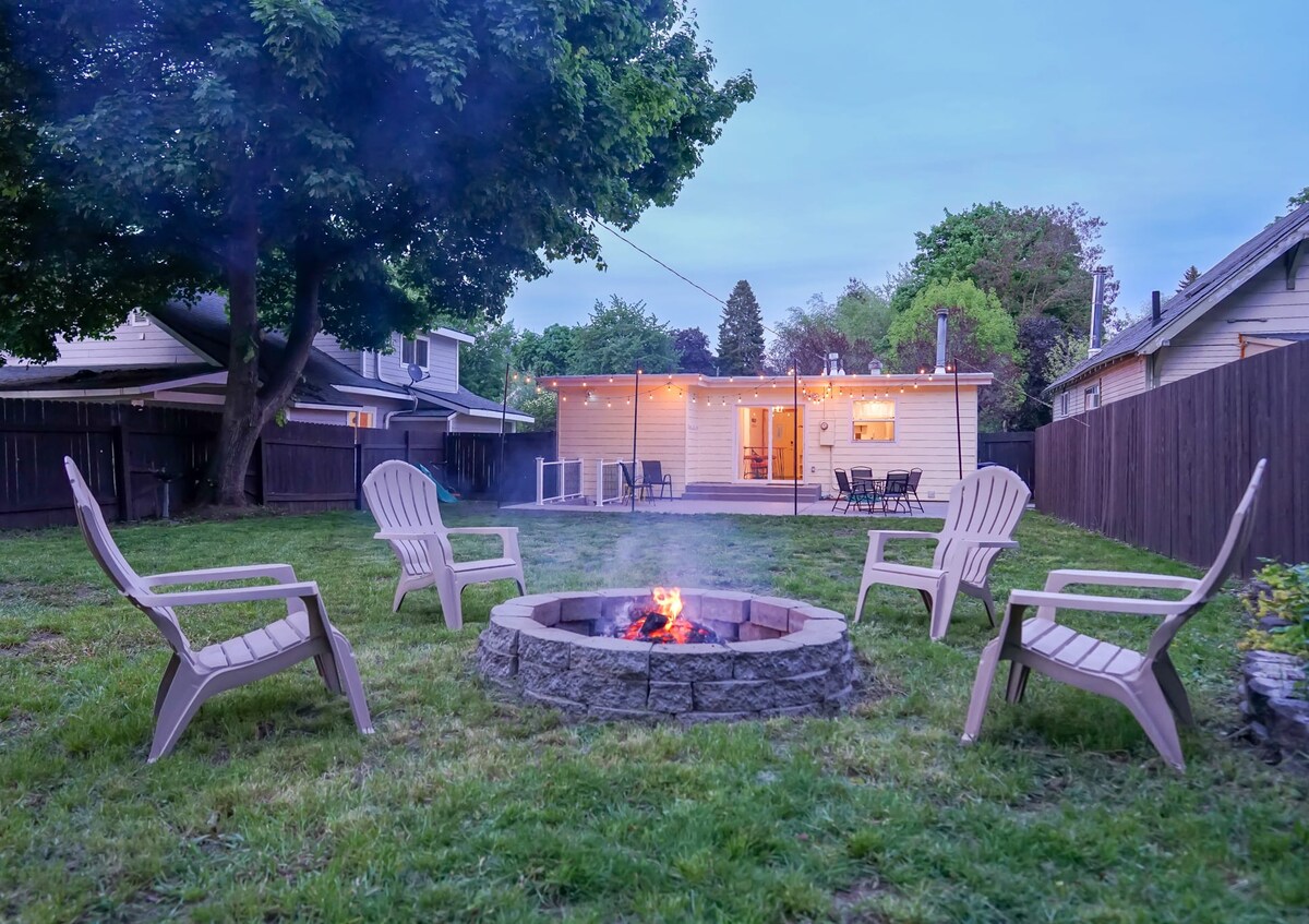 A spacious backyard features a fire pit surrounded by four comfortable chairs. The area is enclosed by a fence for privacy. Soft lighting from string lights highlights the cozy outdoor seating, with the cottage visible in the background.