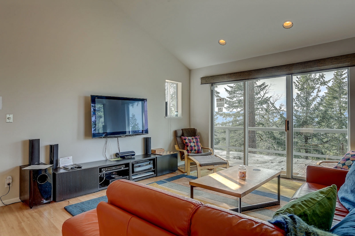 A cozy living room features a large TV mounted on the wall, complemented by a comfortable seating arrangement. Large glass sliding doors provide access to the deck, offering views of surrounding trees. A wooden coffee table sits center stage on a colorful area rug, enhancing the inviting space.