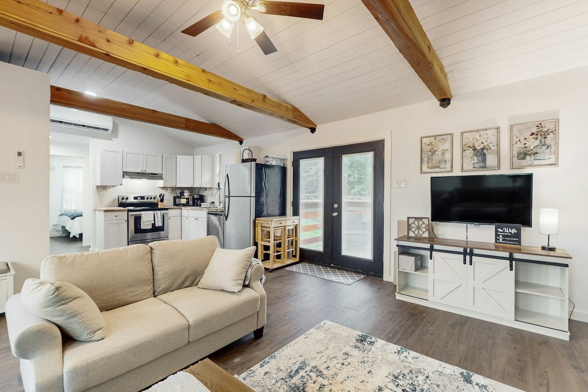 The inviting living area features a comfortable sofa, a wall-mounted television, and light-colored cabinetry. Natural light enters through glass doors leading to an outdoor space, while exposed wooden beams add character to the ceiling.