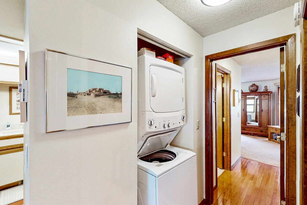 A compact laundry area is seen in a hallway, featuring a stacked washer and dryer for convenience. A framed photograph hangs on the wall, offering a glimpse of the surrounding space. The hardwood floor extends into the entrance of a living area visible in the distance.