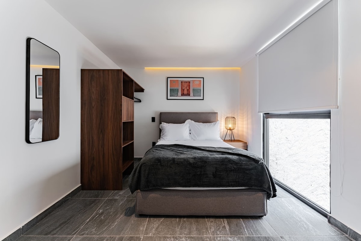 A comfortable sleeping area is featured with a spacious bed, complemented by stylish bedding. A sleek nightstand with an elegant lamp and a contemporary mirror adds to the modern aesthetic. Natural light enters through the large window, accentuated by minimalist window treatments.