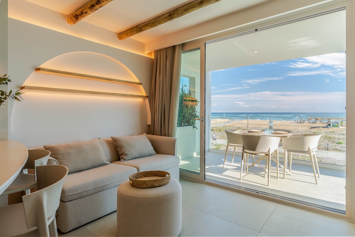 A spacious living area features light-colored furnishings and large glass doors that open to a private balcony. Natural light floods the room, highlighting the minimalist decor. The sea can be seen in the distance, with comfortable outdoor seating set on the balcony.