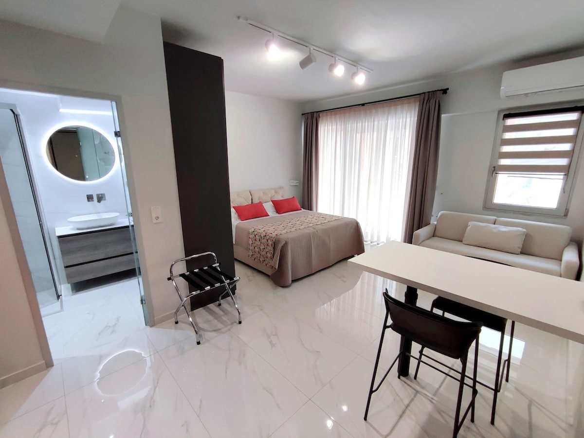 A bright studio space includes a spacious bed with a light-hued coverlet and red accent pillows. A comfortable couch is positioned next to a dining table with four chairs. Natural light filters in through large windows adorned with sheer curtains, enhancing the overall openness of the room.