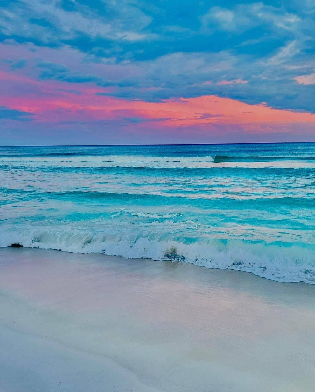 A serene beach scene is presented, featuring gentle waves lapping at the shoreline. The sky displays a gradient of colors, transitioning from soft pink to deep blue, indicating a delightful sunset. The tranquil ocean reflects the serene hues of the evening.