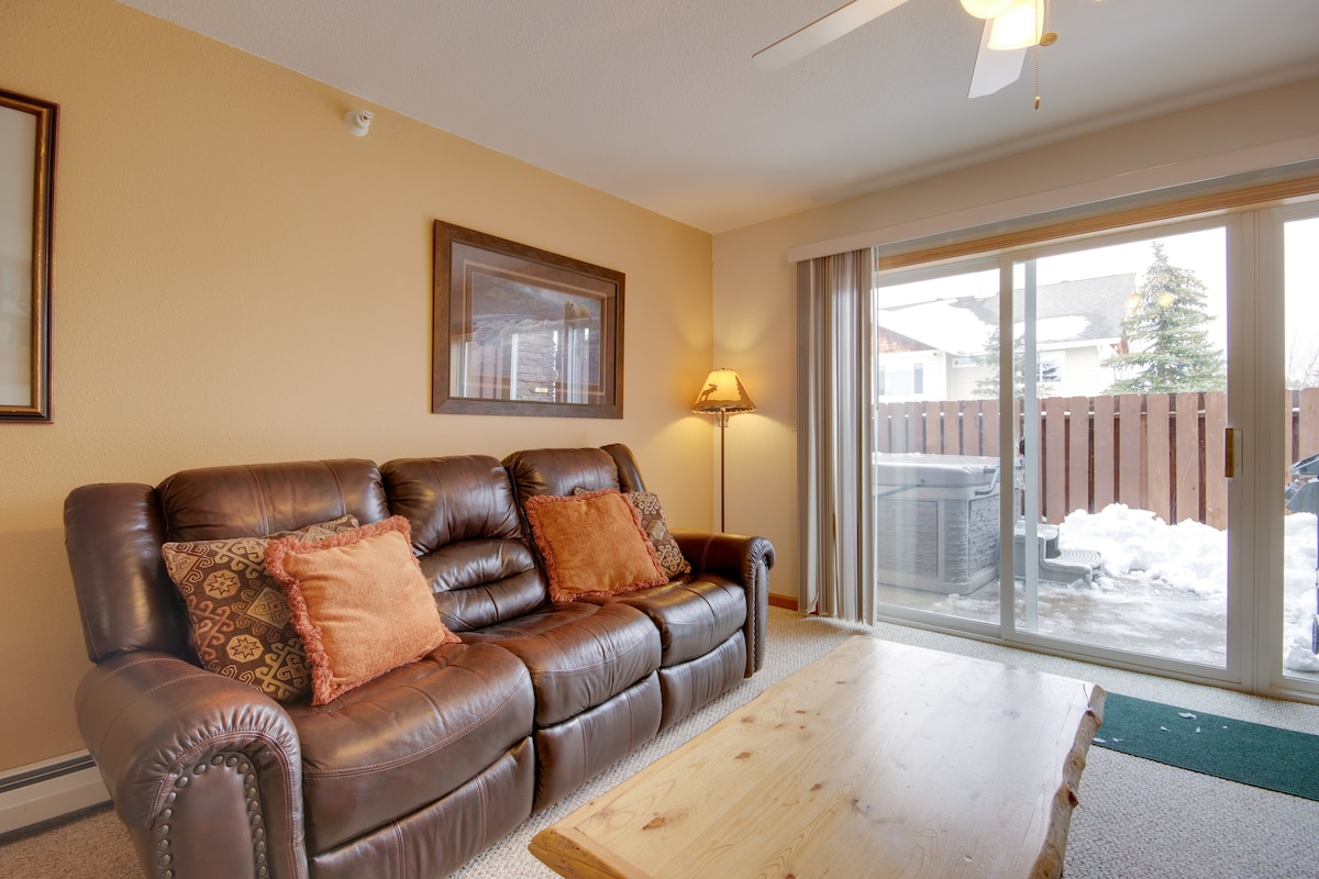A comfortable living area features a leather sofa adorned with decorative cushions. A wooden coffee table provides a functional centerpiece. Natural light streams through sliding glass doors, leading to an outdoor space with a hot tub and views of a fenced area, partially covered in snow.