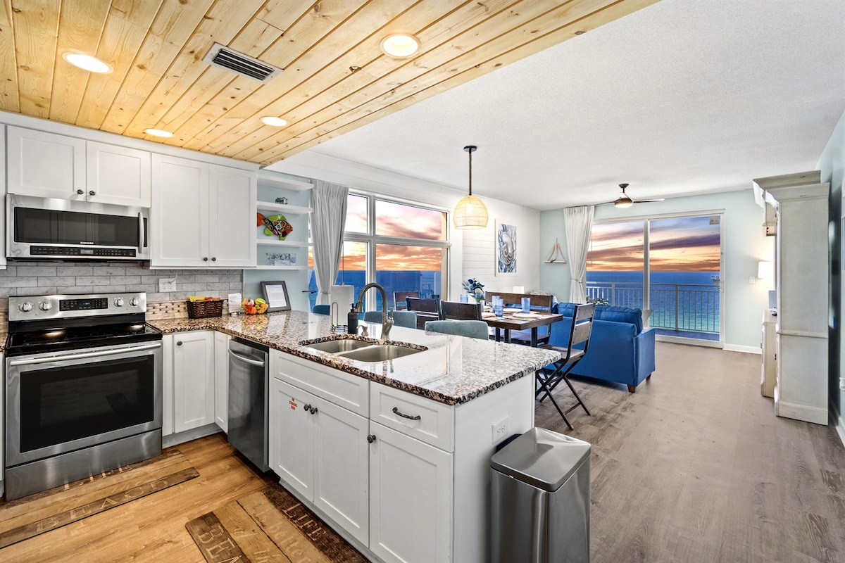 A modern kitchen is equipped with stainless steel appliances and a granite countertop. Open concept living and dining areas showcase large windows revealing stunning ocean views and a colorful sunset. Light wooden beams complement the bright interior design.
