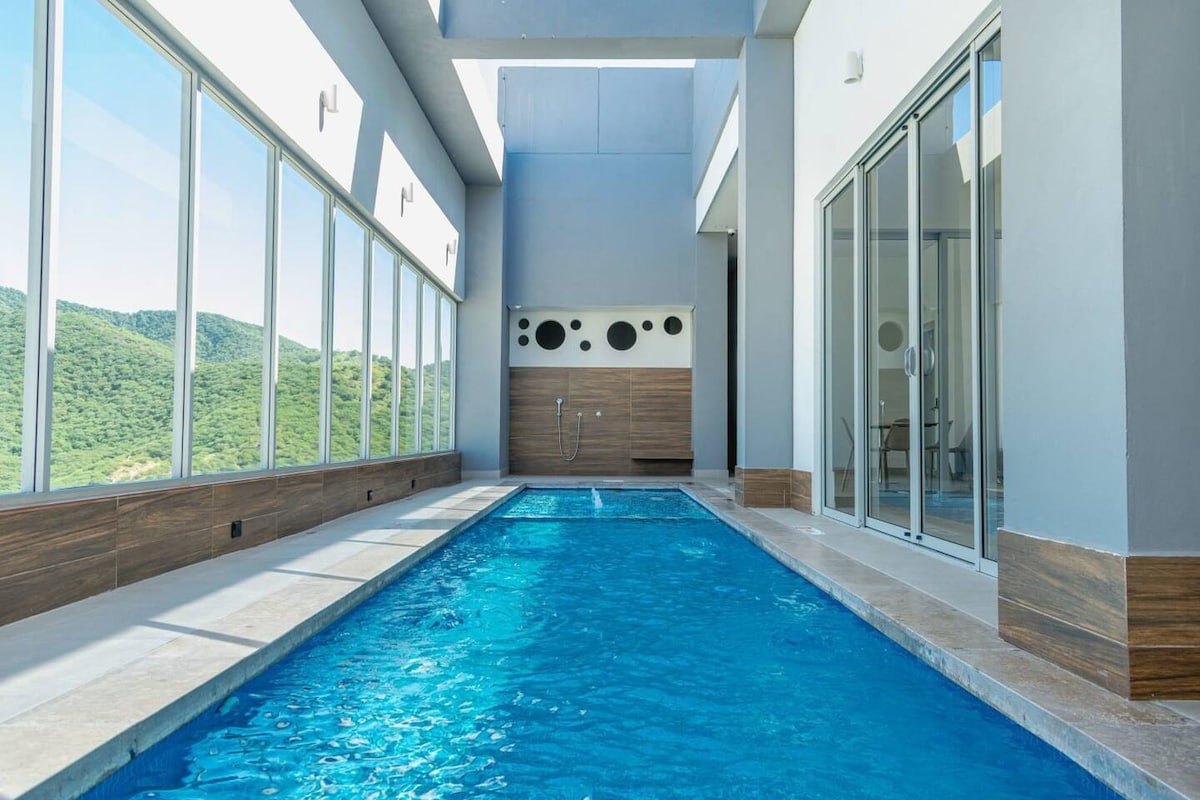 An indoor swimming pool is framed by large glass windows, providing views of the surrounding hills. Natural light floods the space, creating a serene atmosphere. The streamlined design features smooth tile flooring and sleek walls, enhancing the tranquil environment.