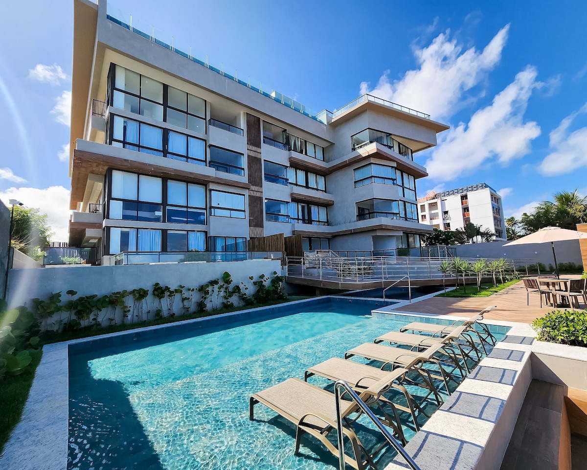 The modern building features multiple levels with large glass windows, allowing natural light indoors. A well-maintained pool is visible in the foreground, accompanied by several lounge chairs arranged on the deck. Lush greenery surrounds the pool area, creating a serene environment.