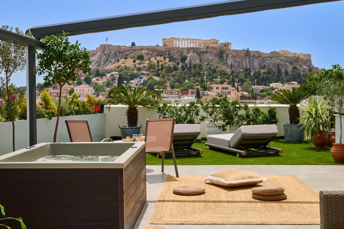 A private terrace features a hot tub surrounded by greenery and comfortable loungers. A neutral-colored rug and cushions are placed on the grass, providing an inviting spot to relax. In the background, the Acropolis is visible against a clear blue sky.