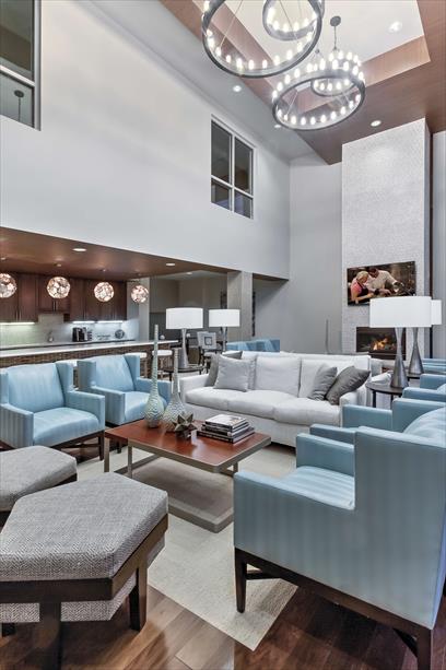 The spacious communal lounge features modern blue chairs and plush seating arranged around a coffee table. A sleek design is highlighted by a high ceiling, elegant light fixtures, and a large television mounted on the wall. A welcoming atmosphere is created by warm wood accents.