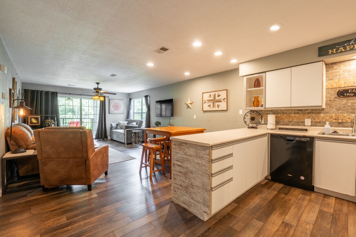 An open concept living space is depicted, featuring a well-equipped kitchen with modern appliances and a breakfast bar. The living area includes a comfortable seating arrangement, a television, and natural light streaming in through large windows that overlook the surrounding greenery.
