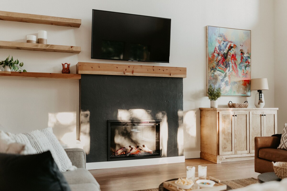 A modern living room features a wall-mounted TV above a sleek electric fireplace. Natural light filters through the space, highlighting wooden shelves with decorative items. A neutral sofa and armchair provide comfortable seating, complemented by a light-colored sideboard displaying artwork and decorative accents.
