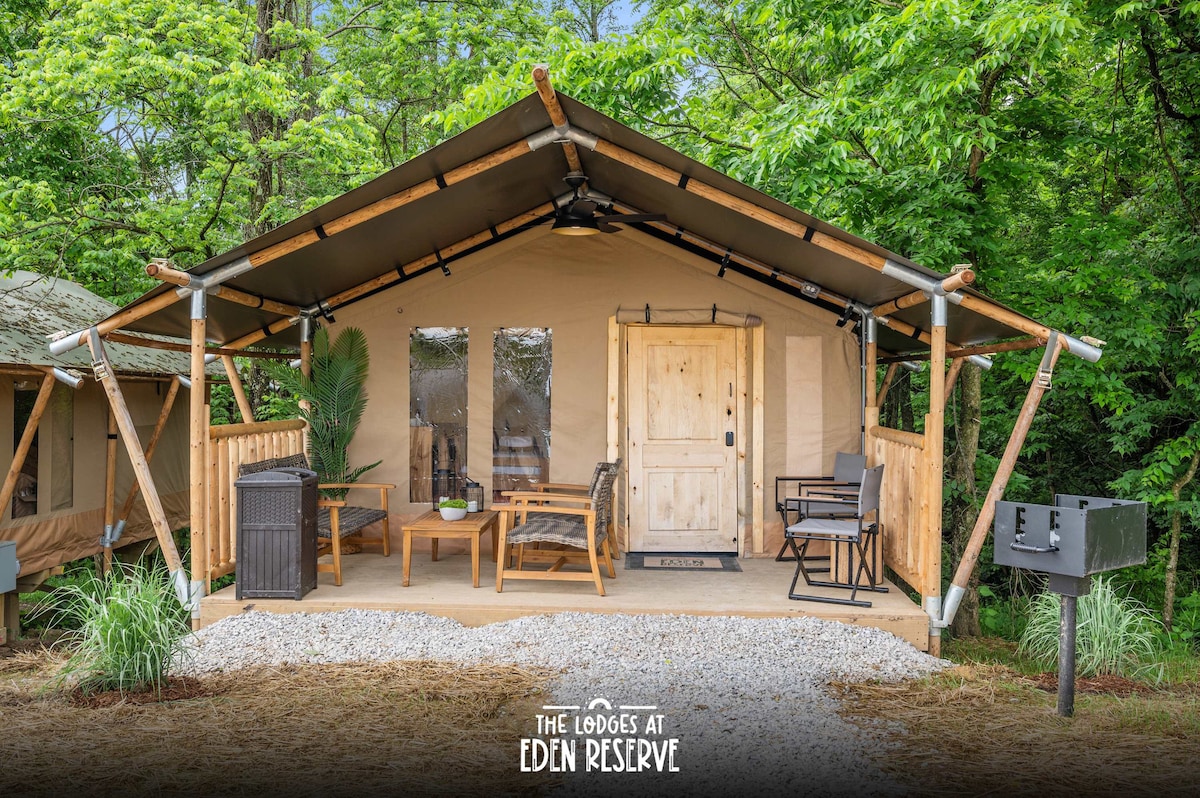 The exterior of a safari-themed tent features a wooden deck furnished with a small dining table and chairs. A charcoal grill is positioned nearby. The entrance displays a wooden door, framed by large windows, surrounded by greenery and gravel pathways.
