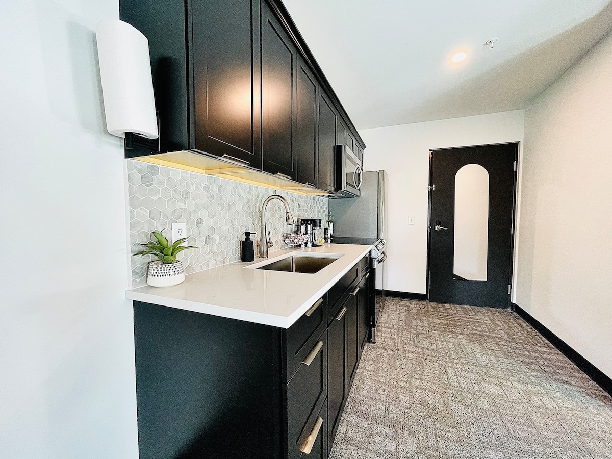 A modern kitchen area features sleek black cabinetry, stainless steel appliances, and a stylish backsplash with hexagonal tiles. The countertop is white and spacious, complemented by an under-cabinet light. A small plant adds a touch of greenery, enhancing the inviting atmosphere.
