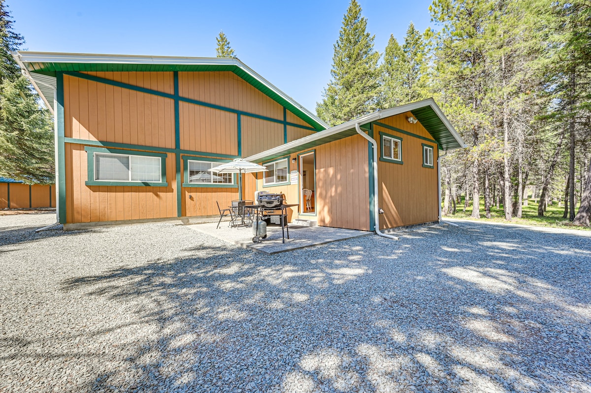 The exterior of the home is set among tall trees, showcasing a combination of orange and green siding. A gas grill is placed on a furnished patio, which features a table and chairs. The gravel area offers ample space for outdoor activities.