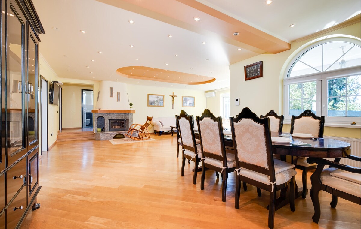 A well-lit dining area features a large wooden table surrounded by elegant chairs. A cozy fireplace is located in the corner, complemented by smooth wooden flooring. Arched windows create a bright atmosphere, showcasing views of the surrounding greenery.