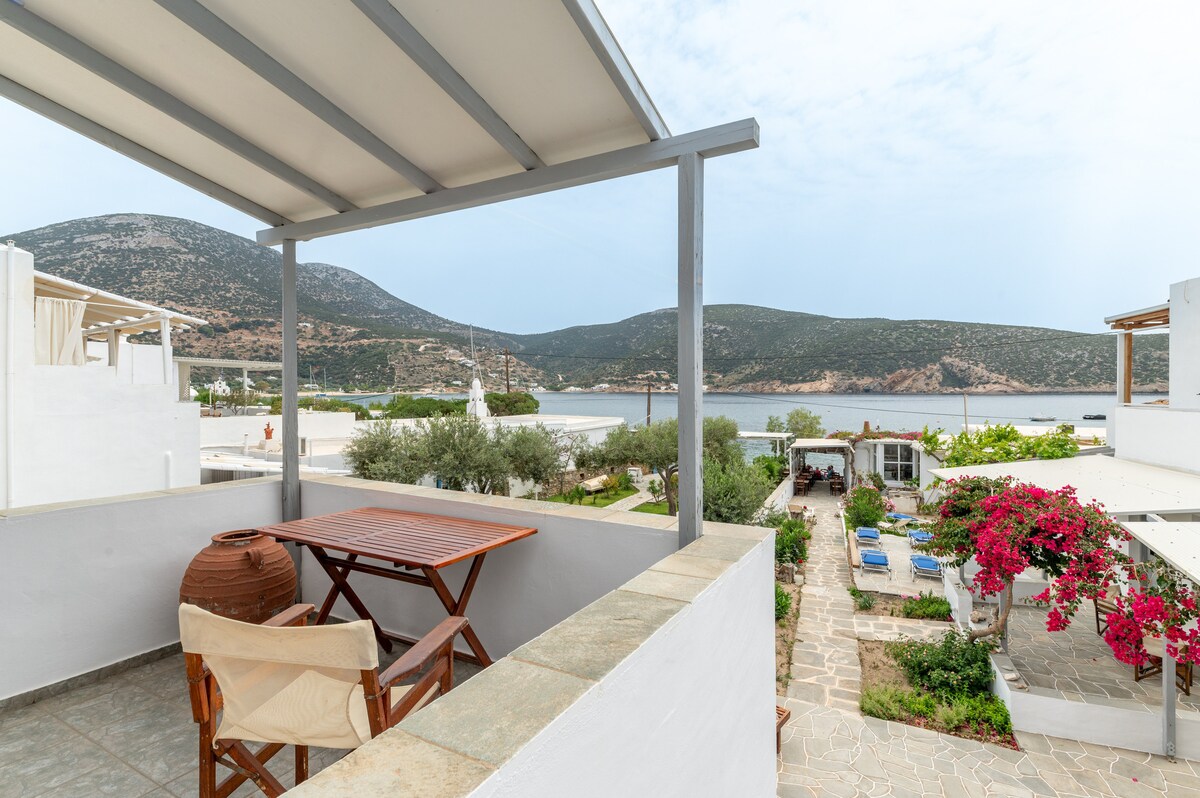 A private terrace offers outdoor seating with a small table and chairs, framed by the surrounding landscape. The view includes the tranquil Aegean Sea and distant hills, while vibrant flowers and greenery enhance the outdoor space.