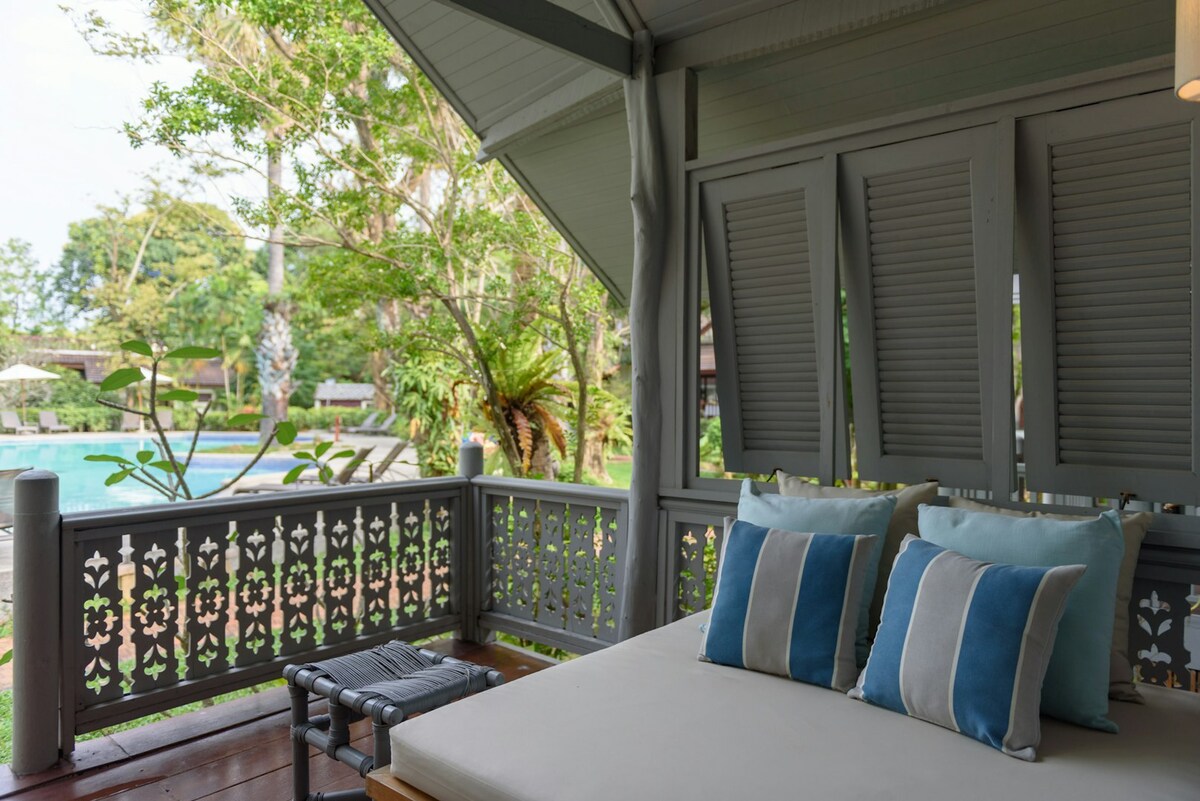 A shaded seating area features a spacious daybed adorned with soft blue and white cushions. Lush greenery surrounds the space, providing a serene view of the nearby swimming pool, framed by tropical plants and trees.