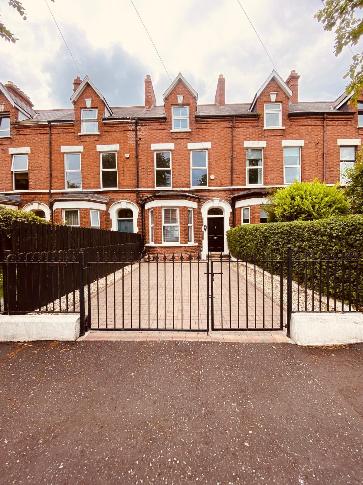A charming Victorian townhouse is enclosed by a black wrought-iron gate, leading to a neat pathway and manicured hedges. The red brick façade features architectural details typical of the period, framed by tall windows on either side.
