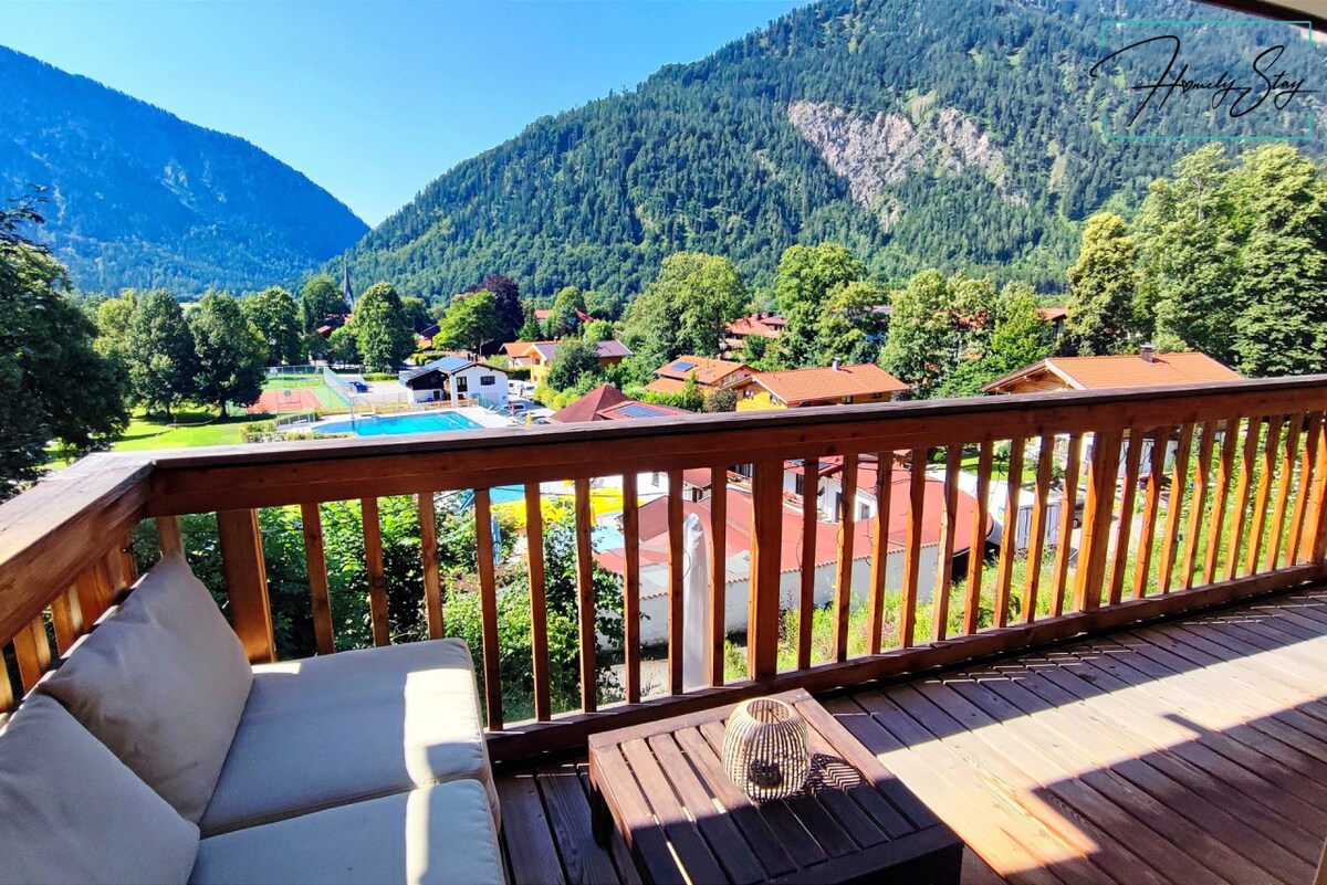 A spacious balcony features comfortable seating, offering expansive views of the surrounding mountains and lush greenery. The landscape includes a glimpse of a swimming pool and nearby charming residences, framed by the rich colors of nature.