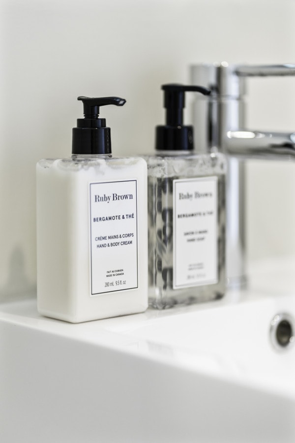 Two elegantly designed Ruby Brown products are displayed on a clean vanity. A creamy hand and body lotion is positioned beside a clear soap dispenser, both featuring minimalist labels. The sink faucet reflects soft light, enhancing the contemporary bathroom ambiance.