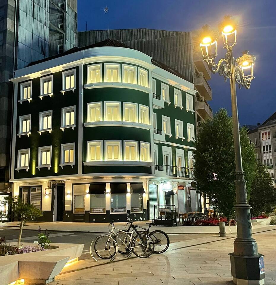 An elegant three-story building featuring a green facade, illuminated windows, and a distinct architectural style. Two bicycles are parked nearby, and outdoor seating is visible. The surroundings include well-maintained pathways and decorative street lamps, contributing to a pleasant urban atmosphere.