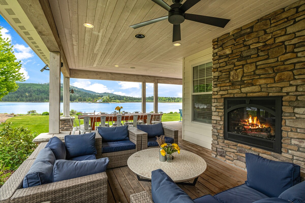 A spacious outdoor seating area features comfortable blue cushions arranged around a circular table. A stone fireplace adds warmth to the covered patio, where views of the waterfront and surrounding greenery can be seen. Natural light enhances the inviting atmosphere.