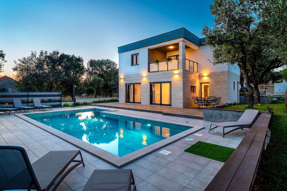 A modern villa features a spacious outdoor area with a rectangular swimming pool, surrounded by lounge chairs. The exterior showcases warm lighting against a clean façade, while a dining table is set on a patio area close to the house, complemented by trees in the background.