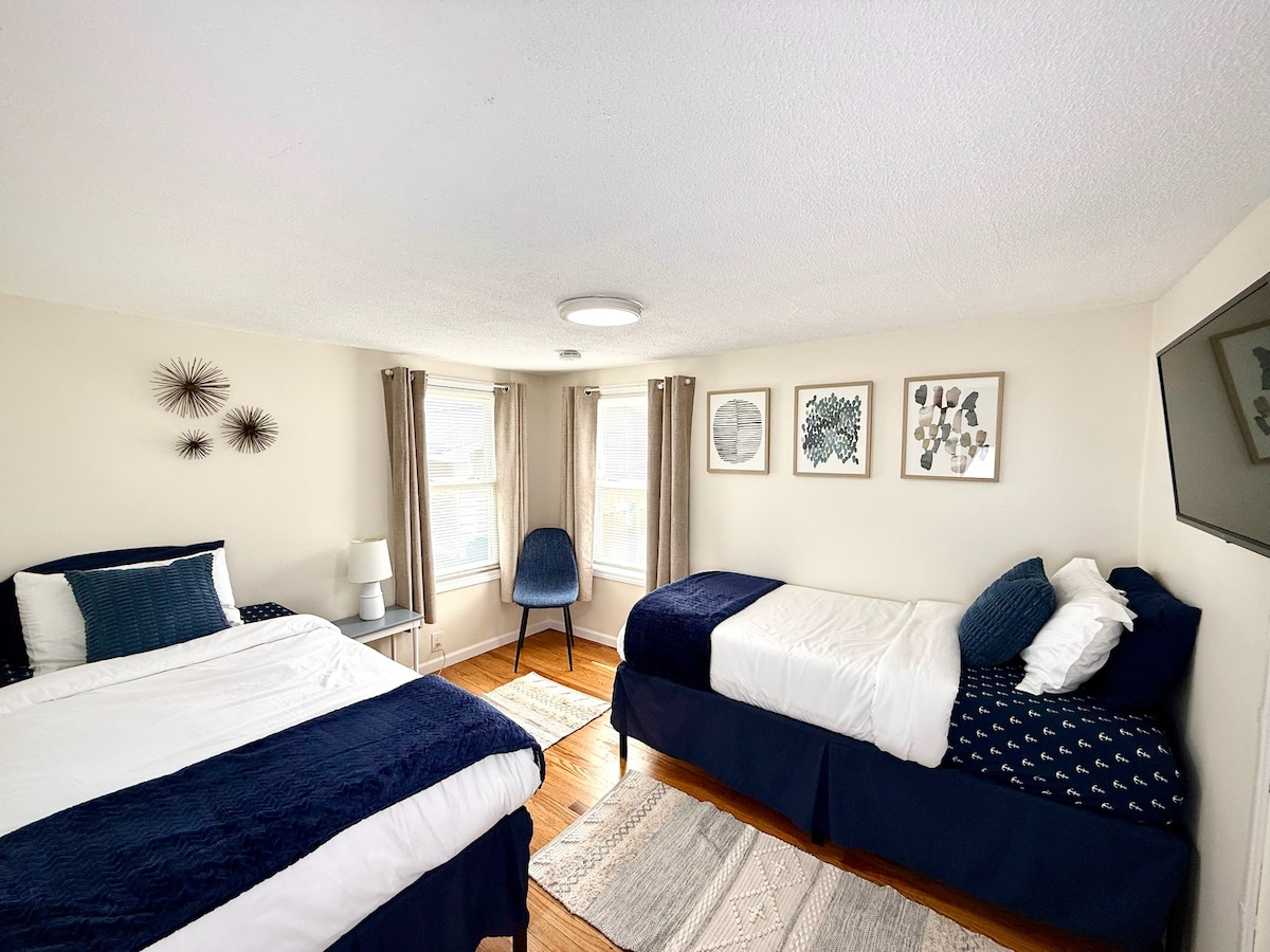 A bright bedroom features two beds dressed in navy and white linens. Natural light fills the space through multiple windows, and wall art adds interest. A small chair is positioned near a television mounted on the wall, enhancing the room's functionality.