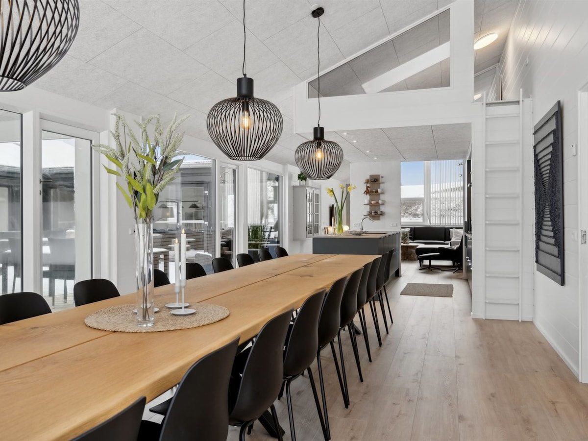 A spacious dining area features a long wooden table surrounded by black chairs. Large windows provide ample natural light, and stylish pendant lights hang from the ceiling. A modern kitchen can be glimpsed in the background, enhancing the open and inviting layout.