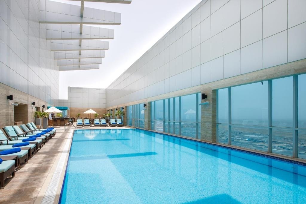 An outdoor pool area is presented, featuring a large swimming pool with clear blue water. Lounge chairs are positioned along the poolside under shaded umbrellas, with floor-to-ceiling windows showcasing views of the surrounding area.