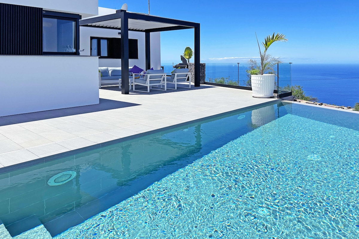 A private heated swimming pool reflects the clear blue sky, bordered by a spacious terrace. Modern seating under a pergola provides shaded relaxation. Ocean views extend beyond the pool, with white walls contrasting against the vibrant landscape.