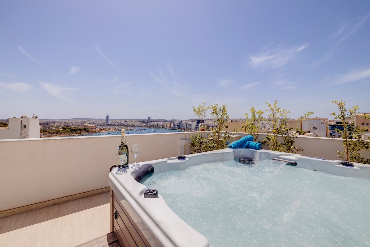 A private rooftop terrace features a luxurious hot tub surrounded by minimal greenery, with stunning views of the harbour and cityscape. A blue towel rests on the edge, alongside two glasses and a bottle of champagne, inviting relaxation and enjoyment in the open air.