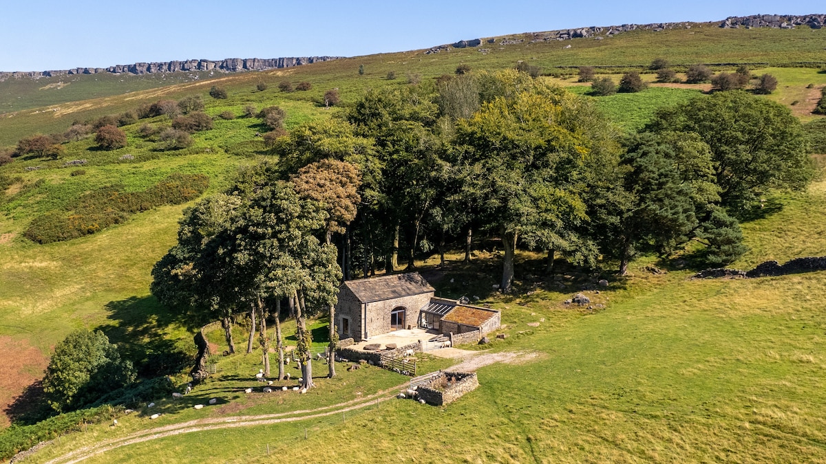An aerial view captures Stanage Barn nestled among ancient trees, surrounded by verdant fields and rolling hills. The structure features a stone exterior and a traditional roof, integrated harmoniously into the natural landscape, offering a secluded escape with scenic views of the countryside.