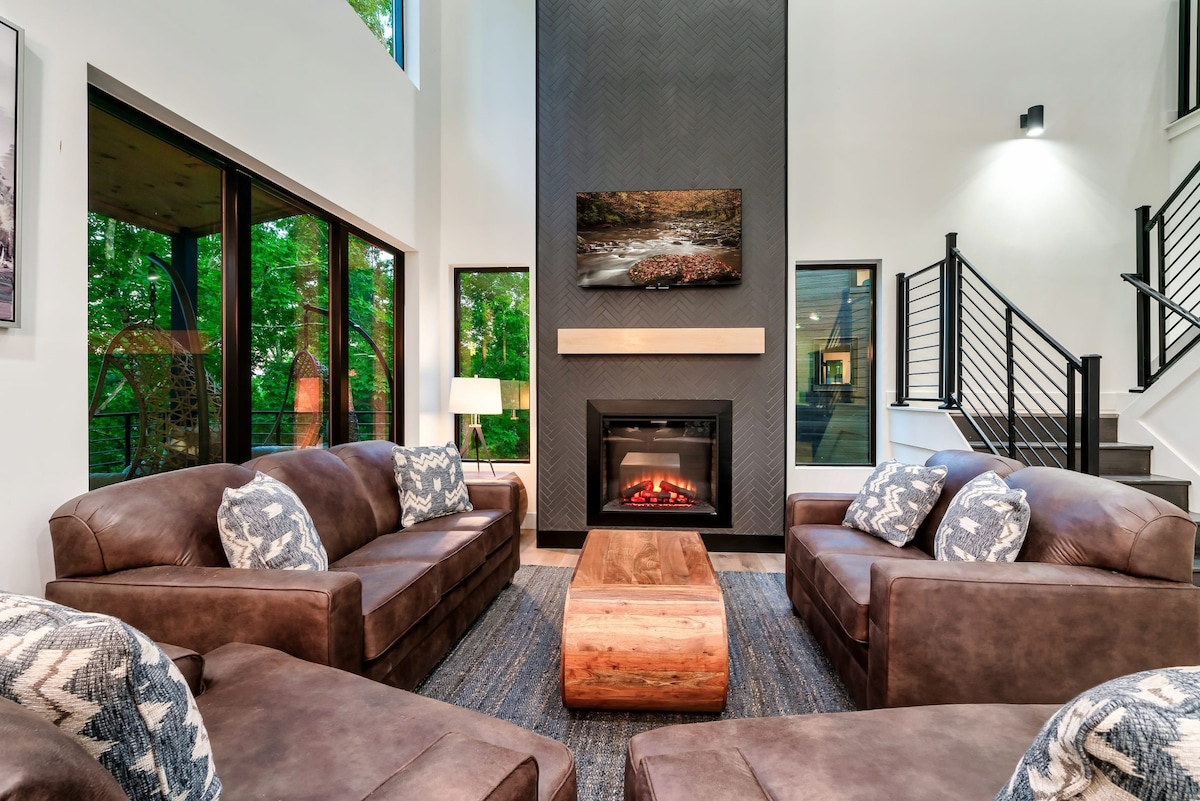 A spacious living area features two leather sofas and a large wooden coffee table. A vertical fireplace is centrally positioned, with a modern art piece above it. Floor-to-ceiling windows allow natural light to illuminate the space, showcasing views of the surrounding greenery.
