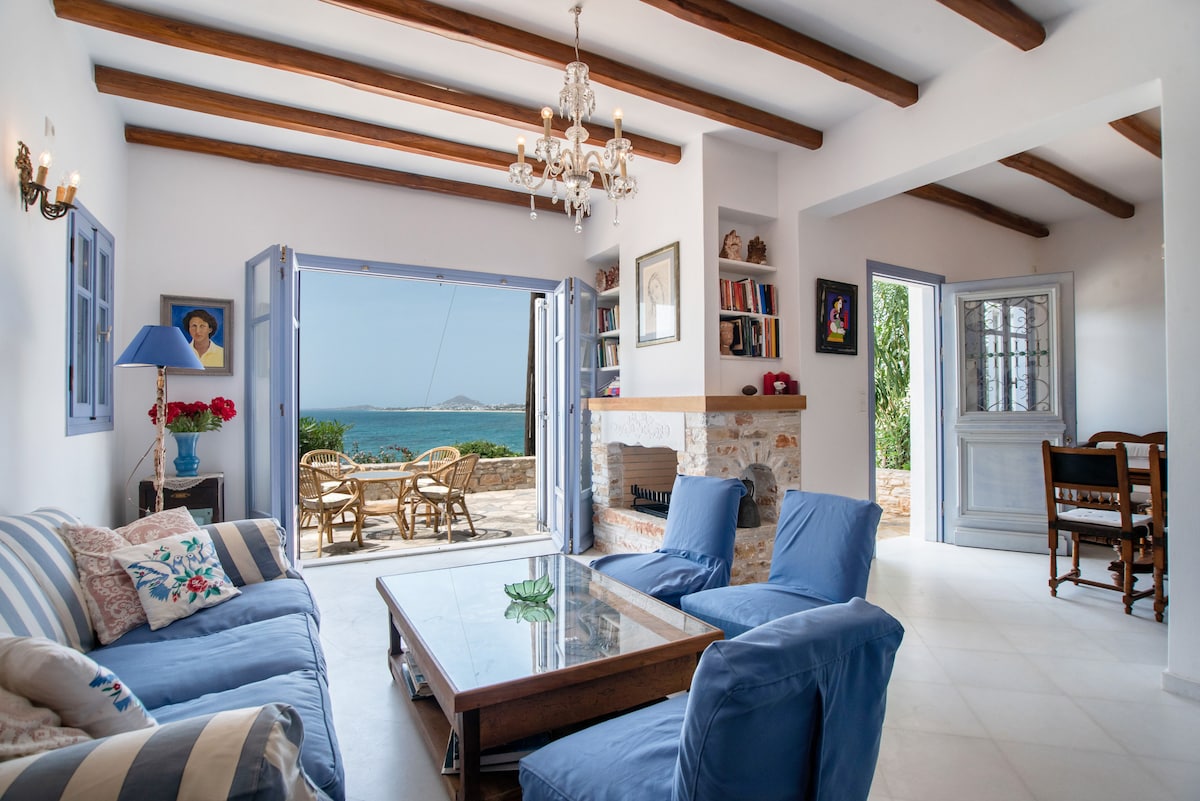 A bright living area is furnished with blue sofas and a glass coffee table. Natural light fills the space through open double doors, revealing a scenic sea view. Bookshelves line the walls, and a dining set is visible just outside, blending indoor comfort with outdoor beauty.