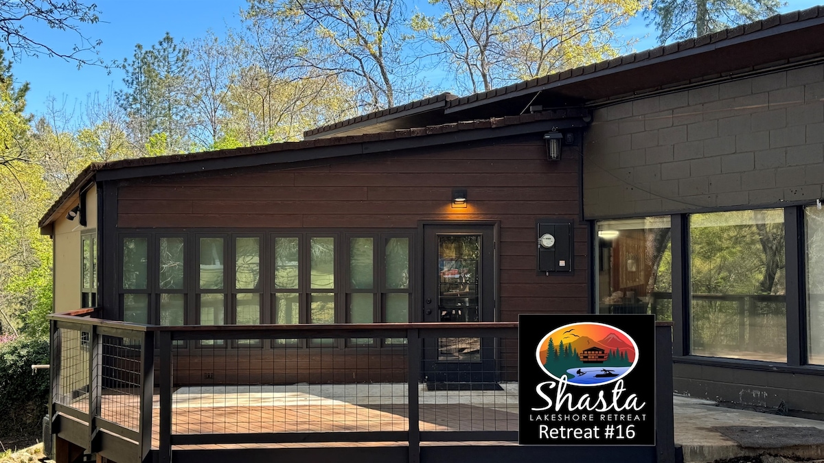 The exterior of Shasta Lakeshore Retreat #16 features a modern wooden structure with large windows, allowing natural light to fill the space. A spacious deck extends from the entrance, bordered by a railing, and surrounded by mature trees, enhancing the serene forest setting.