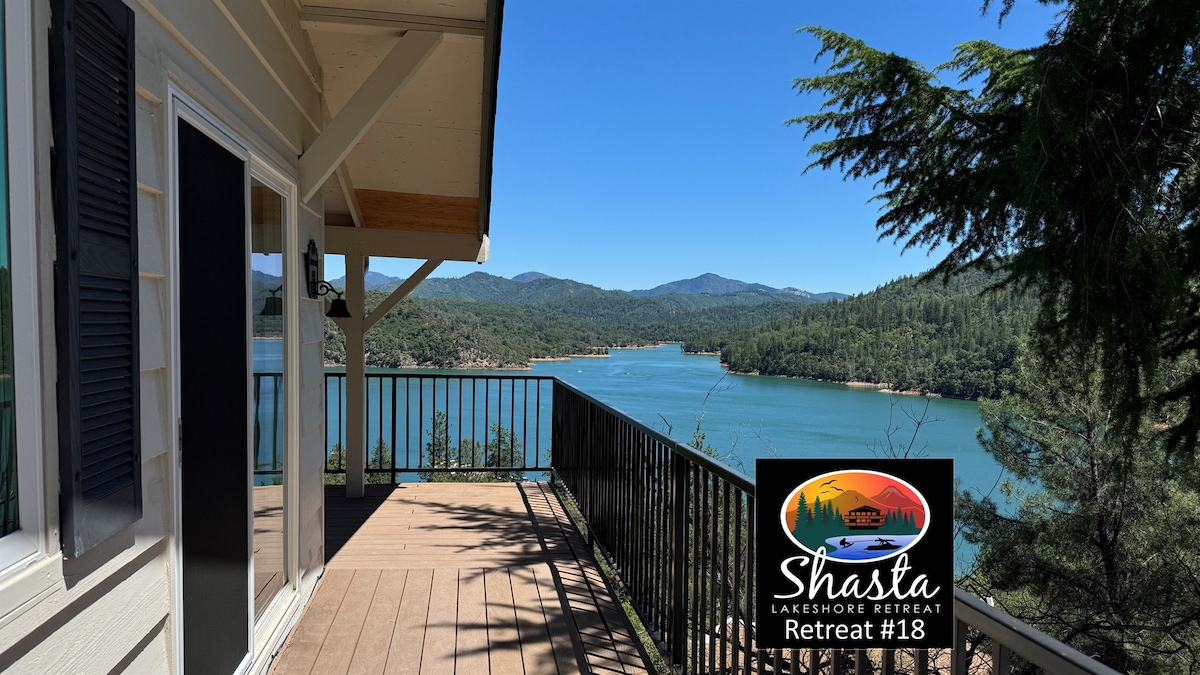 A scenic view from a spacious deck overlooks Shasta Lake, surrounded by lush green hills and distant mountains. The wooden deck features a railing, allowing guests to enjoy the natural landscape and serene water below.