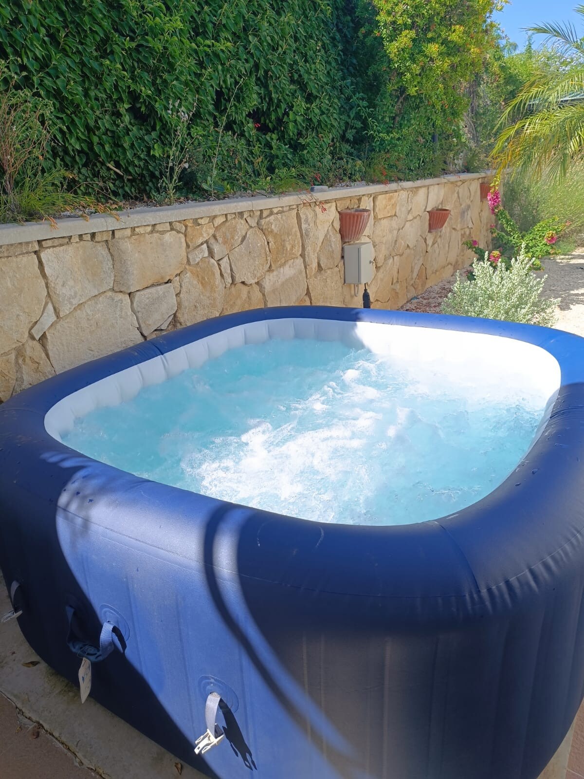 A hot tub is positioned against a stone wall, surrounded by vibrant greenery. The water is gently bubbling, creating a relaxing atmosphere. Sunlight filters through leaves, illuminating the area and enhancing the inviting appearance of the outdoor space.