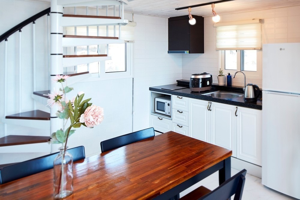 A modern kitchen area is present, featuring a black countertop with essential appliances including a microwave and kettle. A wooden dining table is accompanied by black chairs. Natural light filters through the windows, enhancing the bright atmosphere of the space.