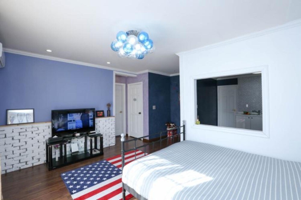 A well-lit room features a spacious bed covered with a striped blanket. A wall-mounted television sits on a stand, and a colorful rug with an American flag design adds a vibrant touch. The room is painted in soft blue tones and has a window view toward the kitchenette.