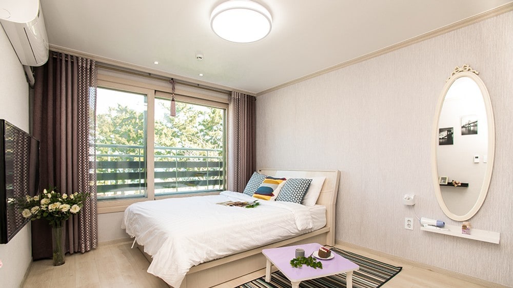 A bright and airy room features a spacious bed adorned with various cushions. A large window allows natural light to fill the space, while decorative curtains frame the view. A round mirror and a small table with greenery add a touch of style to the room.