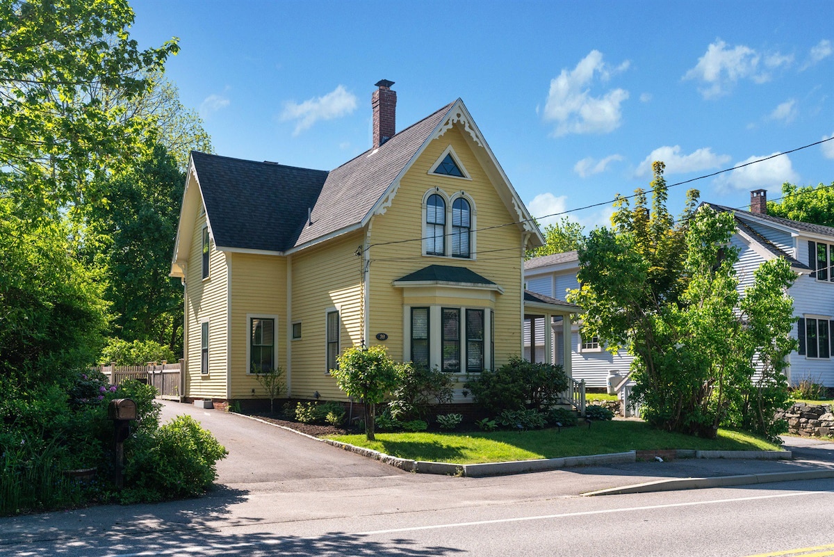 A charming yellow house is prominently featured with a steeply pitched roof and decorative gables. Lush greenery surrounds the property, adding to its inviting presence. Clear blue skies provide a bright backdrop, while the structure showcases multiple windows that enhance its exterior appeal.