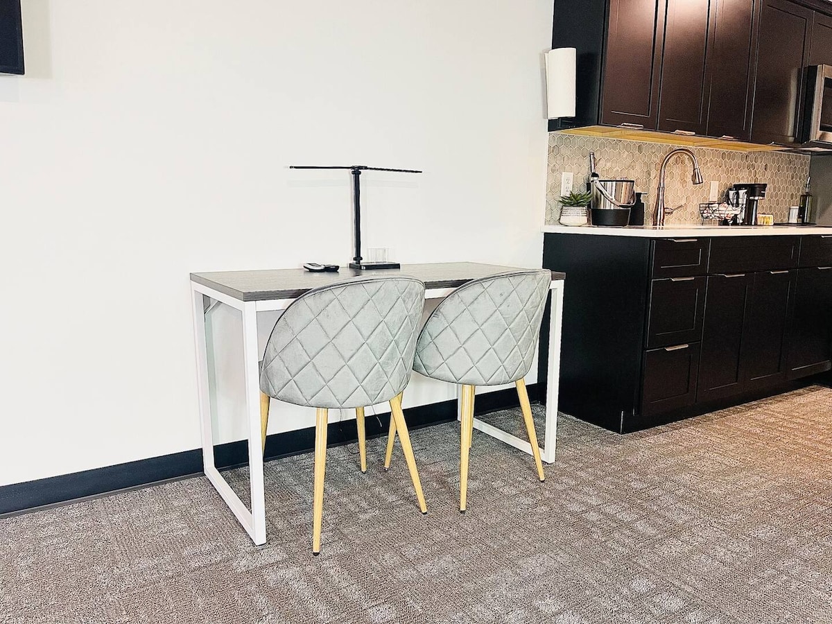 A modern workspace is presented with a minimalist table paired with two upholstered chairs displaying a diamond pattern. A sleek lamp sits on the table, providing illumination. The backdrop features dark cabinetry, enhancing the space's contemporary design.