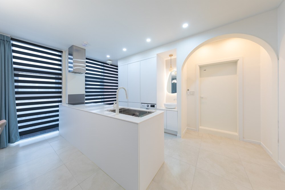A modern kitchen is presented with sleek cabinetry and a central island. Large windows with striped blinds allow natural light to brighten the space. Stainless steel appliances are integrated, complementing the minimalist design, while a dining area is subtly defined in the corner.