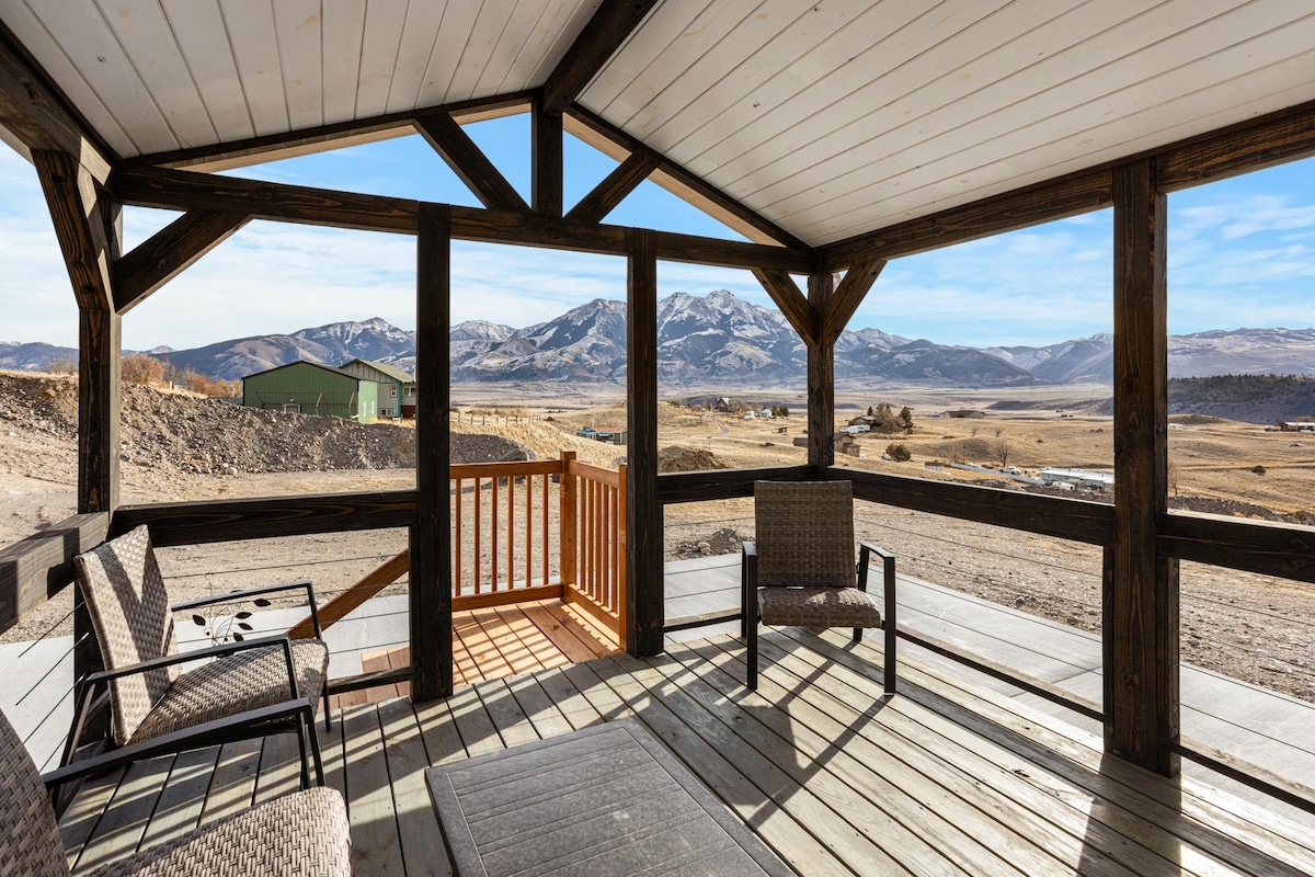 An outdoor deck offers an unobstructed view of the surrounding landscape, featuring mountains and open fields. Two comfortable chairs are positioned around a low table, with wooden beams supporting the structure above. A wooden railing adds a touch of safety while enhancing the view.