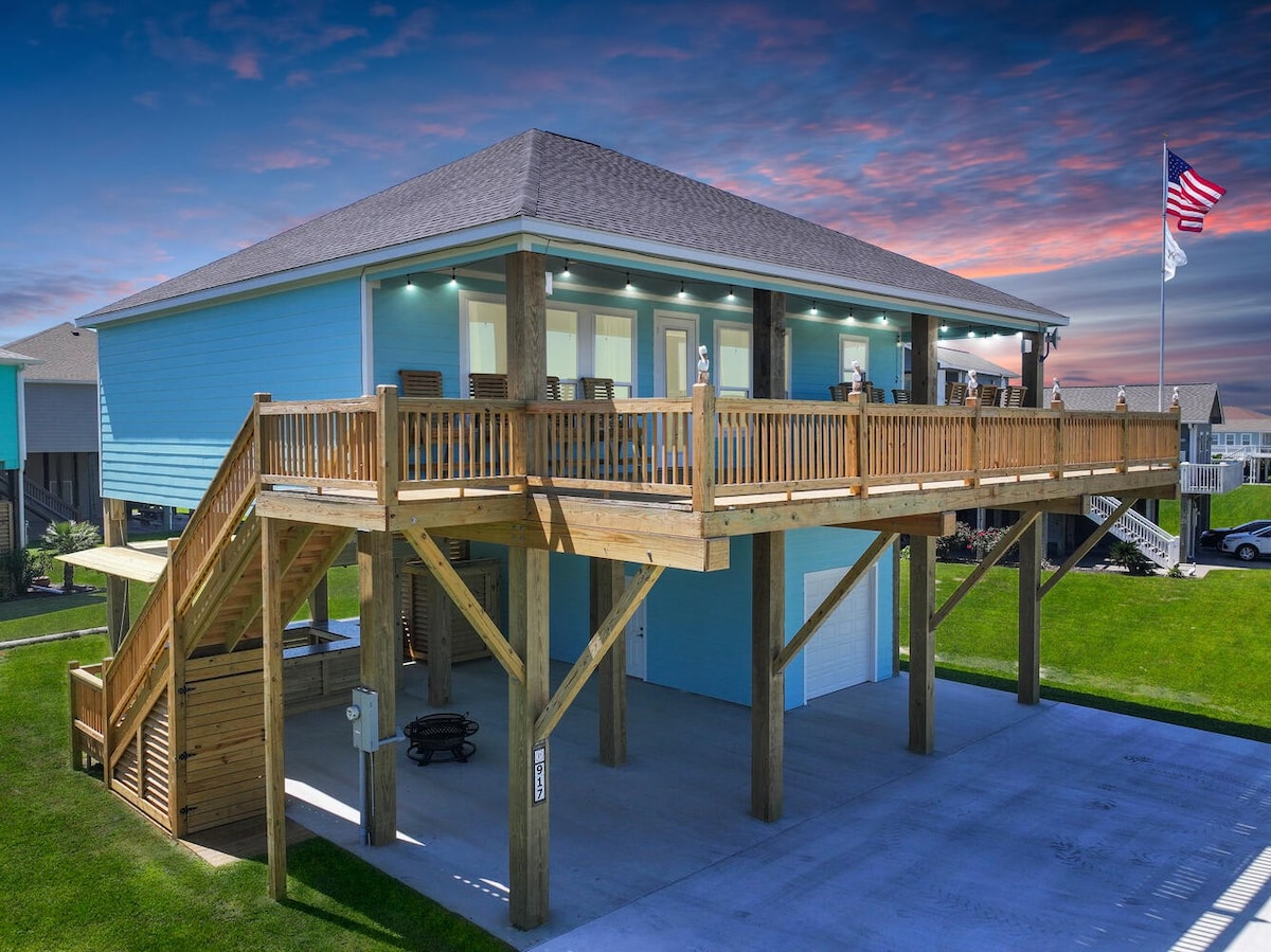 A spacious beachfront house is elevated on stilts, showcasing a large wooden deck adorned with seating. The structure is painted in a soft blue hue, complemented by a gray roof, while an American flag gently flutters nearby, against a colorful sunset sky.