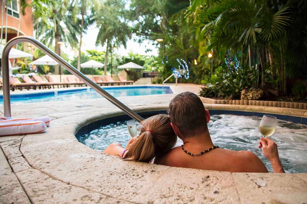 A hot tub is surrounded by lush greenery and palm trees, creating a serene atmosphere. The area features lounge chairs under umbrellas in the background. Two individuals can be seen relaxing together, enjoying drinks while partially submerged in the bubbling water.