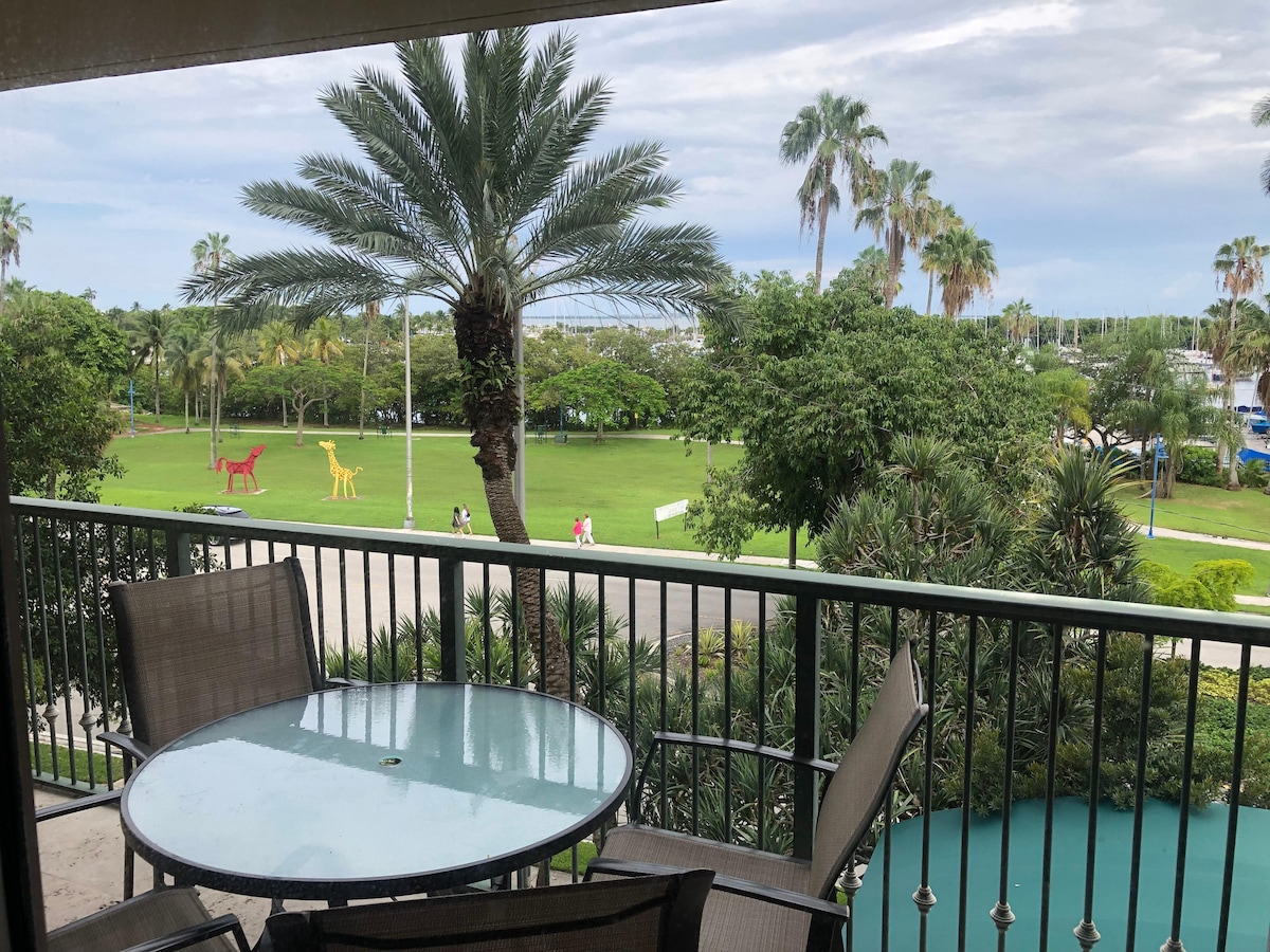 A balcony view is provided, showcasing lush green lawns and palm trees. Colorful sculptures resembling dinosaurs stand prominently on the grounds, while guests stroll along paths in the distance. A glass table with metal chairs offers a comfortable outdoor seating area.