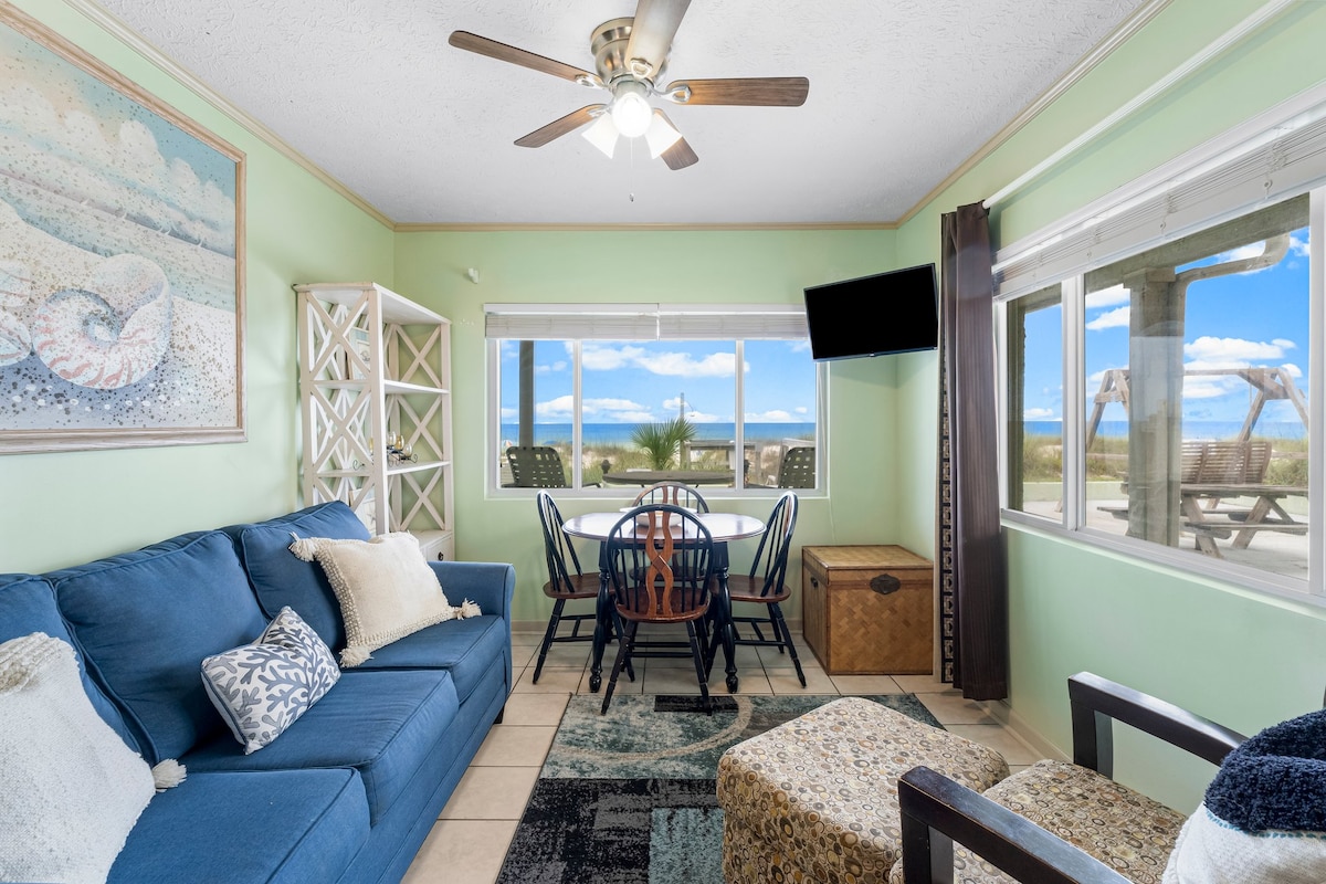 A comfortable living area features a blue sofa with decorative pillows, a dining table with six chairs, and large windows offering expansive views of the beach. Light wood accents and a ceiling fan complement the space. Natural light fills the room, contributing to a relaxed atmosphere.
